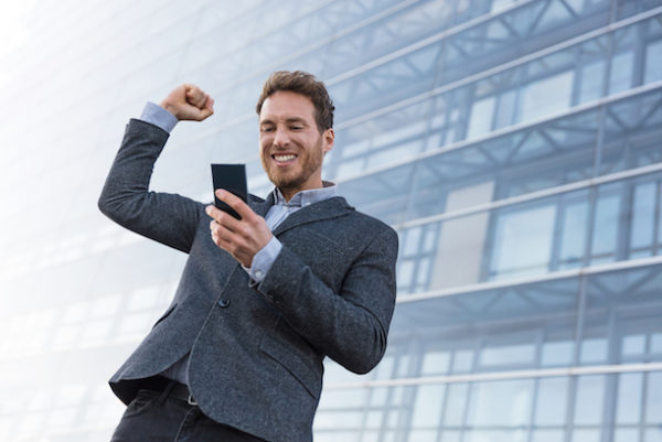a business man looking happily at his cellphone, office building in the background