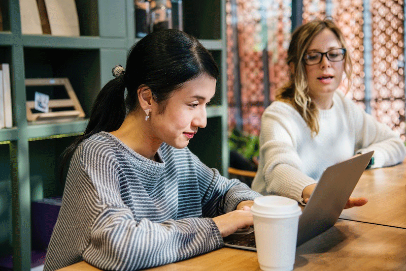 Young woman staring intently at her work laptop, another woman looks on in the background